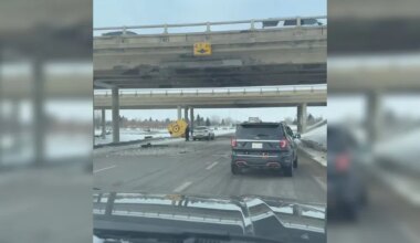 The view from a car travelling on Highway 11 with a damaged overpass overhead. Another vehicle is in front and some equipment parts are in the median ditch.