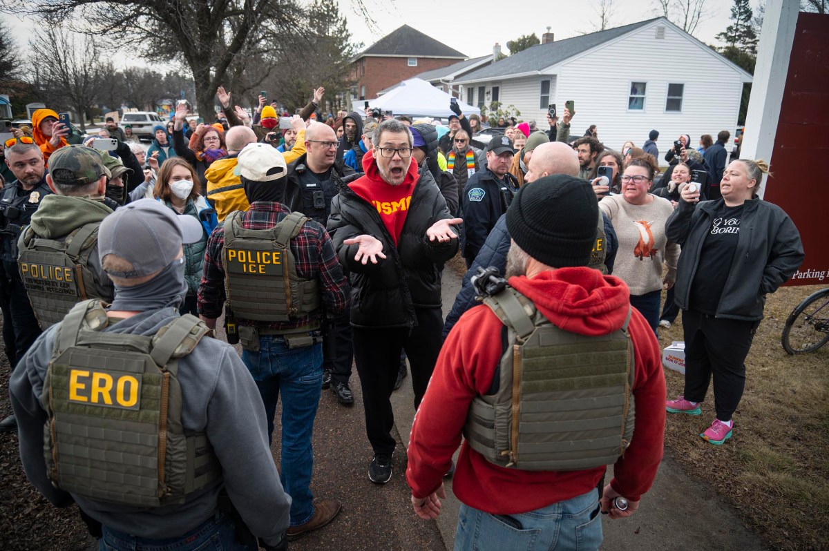 A group of people confront police and ICE officers wearing tactical vests during a tense outdoor encounter in a residential neighborhood.