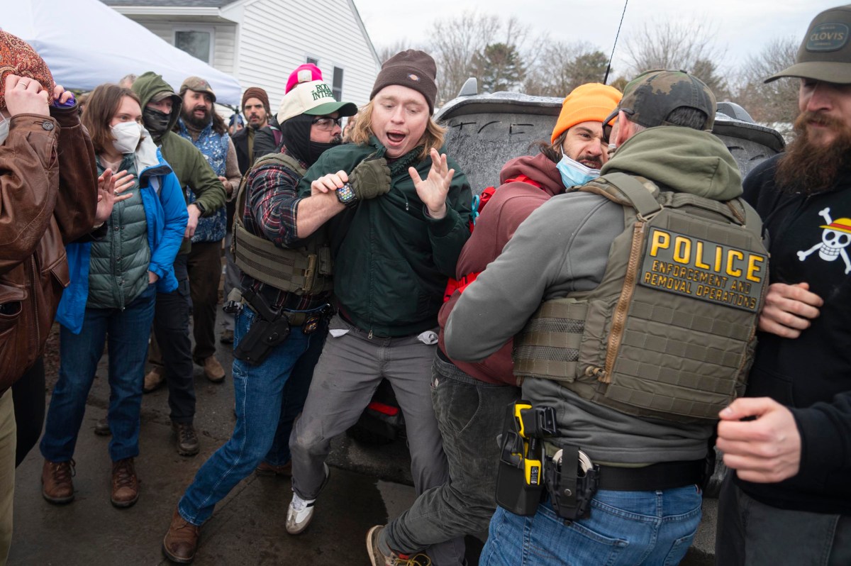 A group of law enforcement officers detain a man in a crowd outdoors; bystanders stand nearby, some wearing masks and winter clothing.
