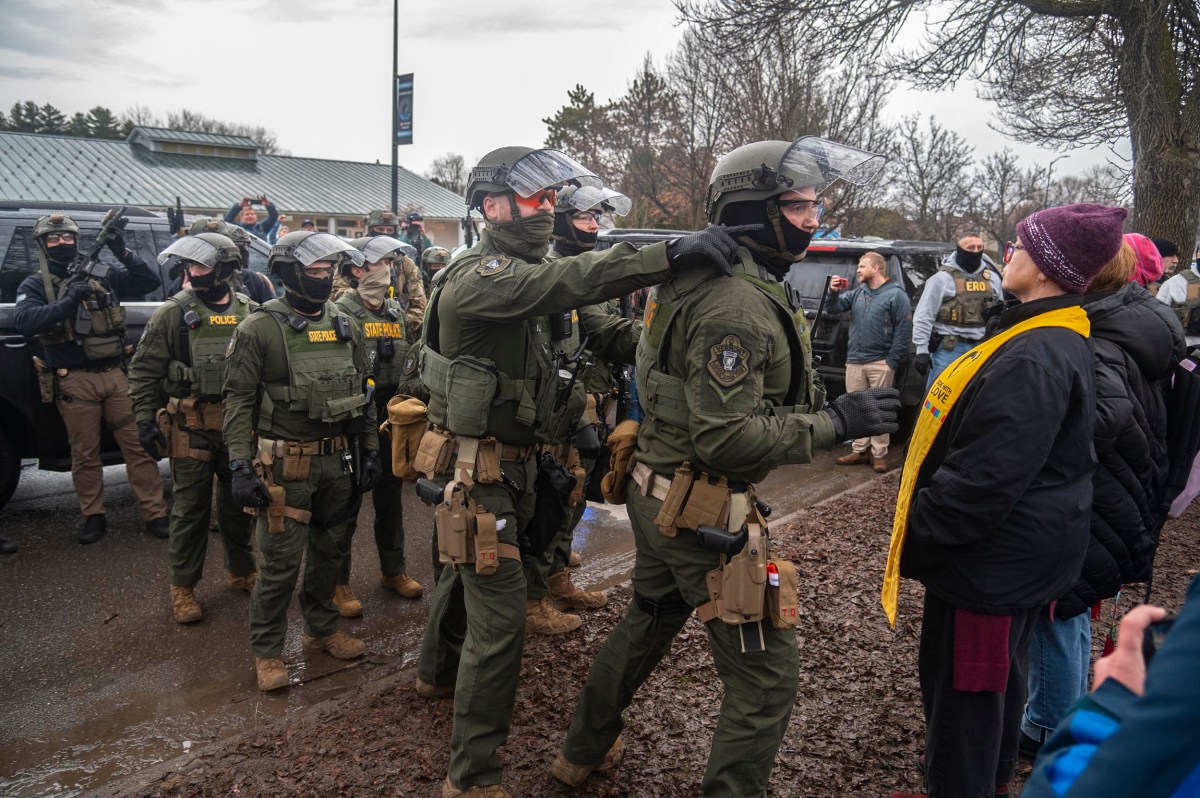 Police officers in riot gear stand in a line facing a group of protesters outdoors; one officer appears to push a colleague forward toward the crowd.