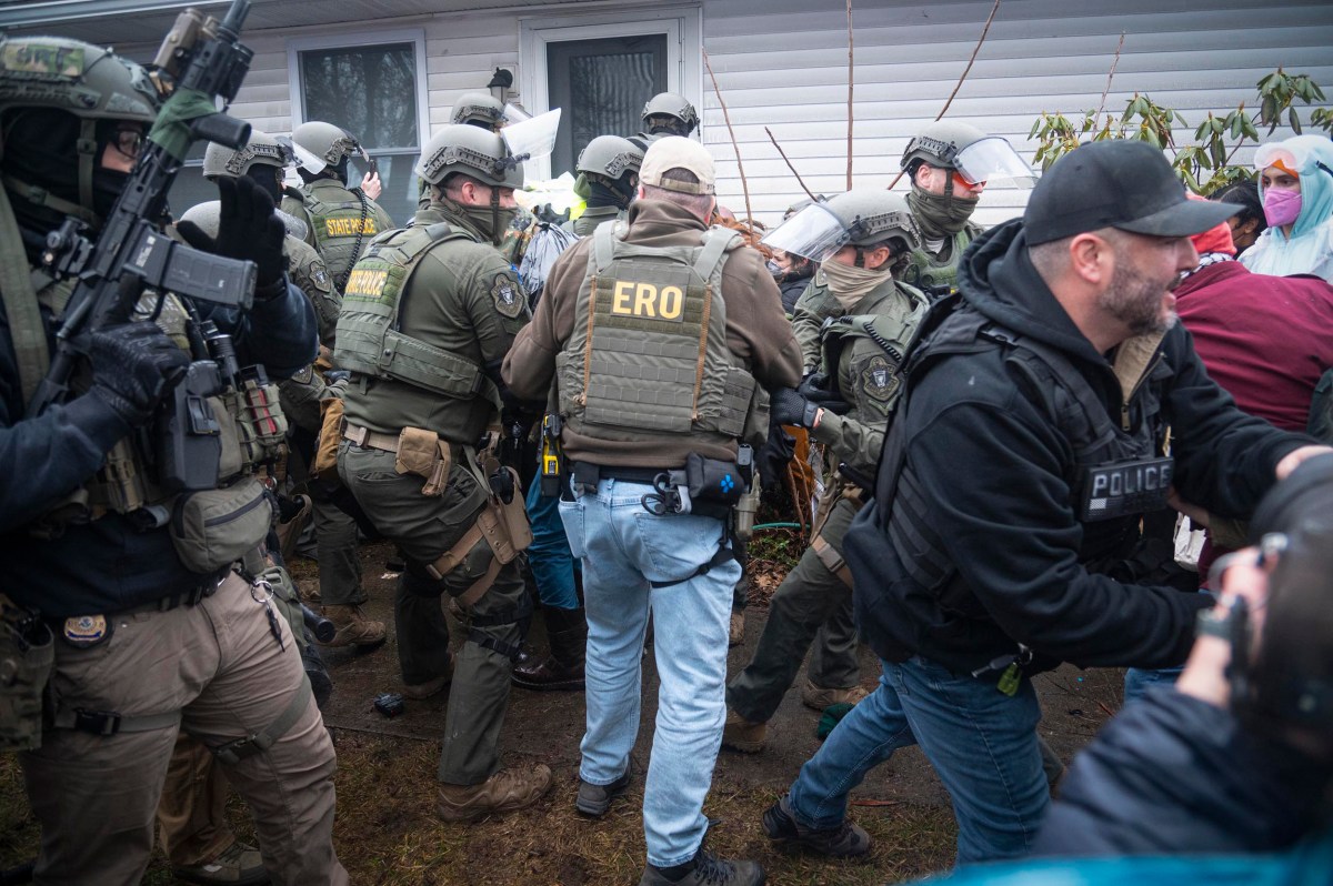 Law enforcement officers in tactical gear gather near the entrance of a house during a daytime operation. Some officers have "ERO" and "POLICE" vests visible.