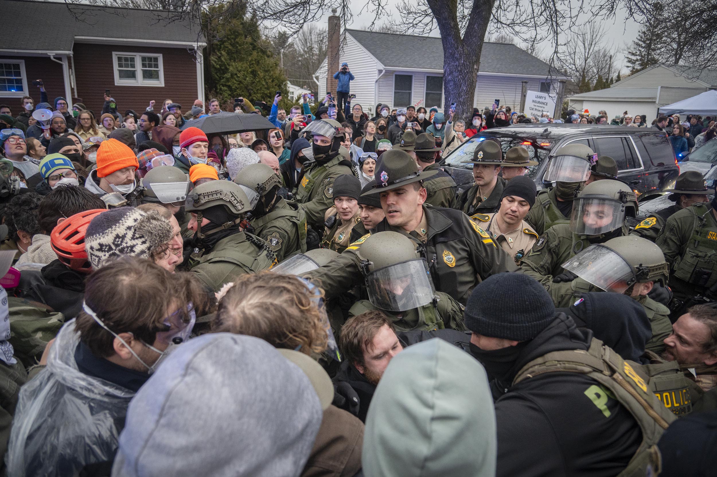 Police officers in riot gear stand in a line facing a dense crowd of protesters on a residential street, with bystanders watching from behind.