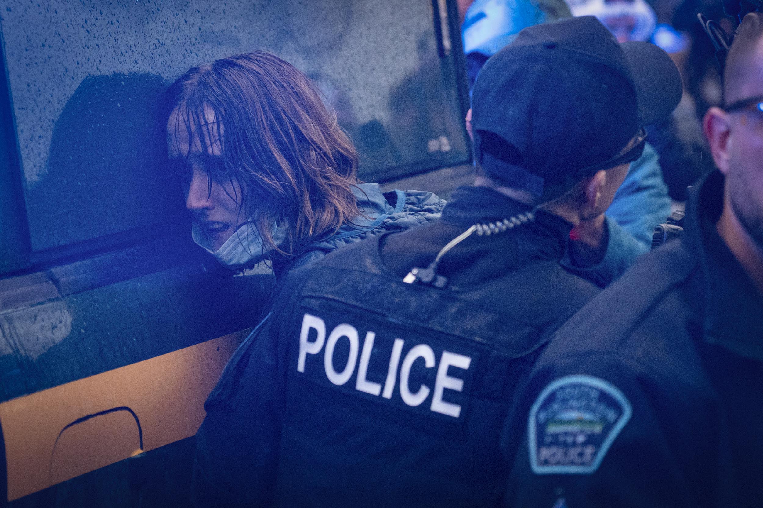 A police officer detains a person by pressing them against the side of a vehicle during a rainy outdoor event.