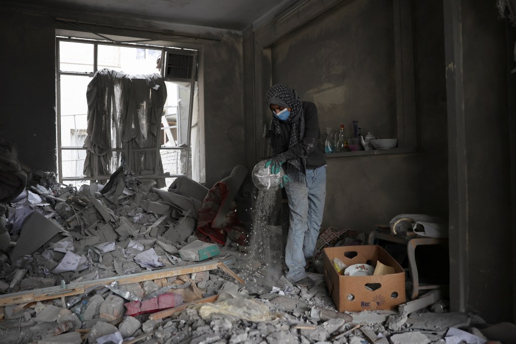 A person wearing a head covering, face mask, and gloves sifts through debris in a damaged room in Tehran.