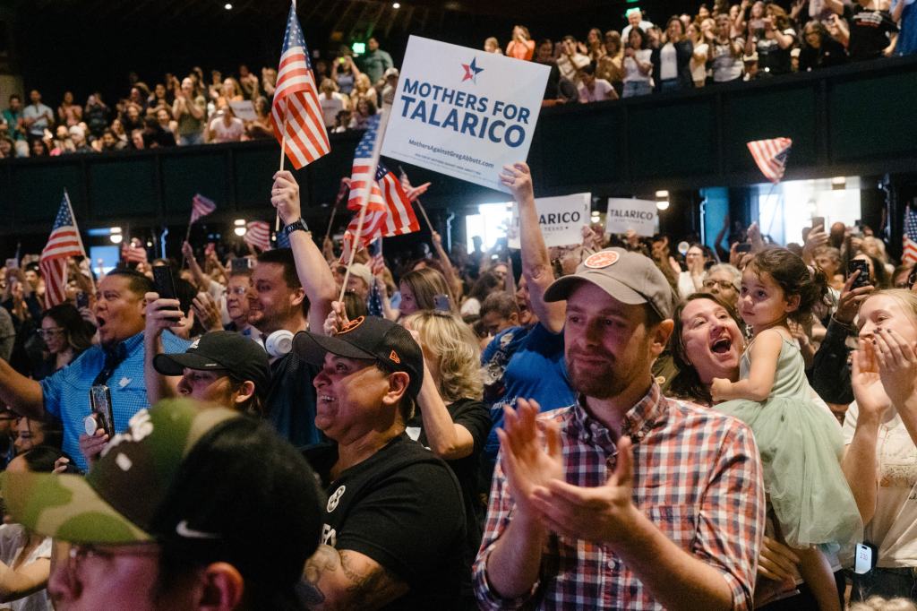 A crowd of supporters clap and smile, holding U.S. flags and signs.