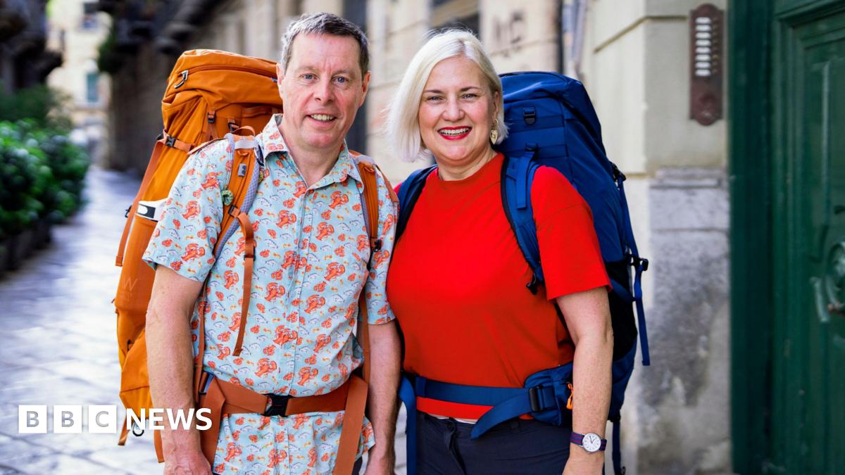 Mark Blythen, in his 60s, who has short grey hair and is wearing a short sleeved blue shirt with a lobster pattern and a large orange backpack, stands next to Margo Oakley, who has short blonde hair, a red t-shirt and a large blue backpack.