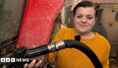 Olly Harrison has short brown hair, he is wearing a red hooded top and a blue jacket and is looking at the camera while stood in front of large stock of fertiliser