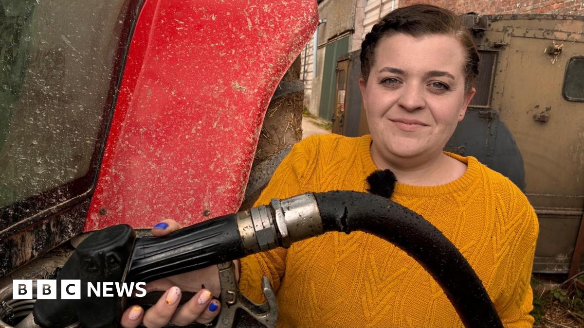 Olly Harrison has short brown hair, he is wearing a red hooded top and a blue jacket and is looking at the camera while stood in front of large stock of fertiliser