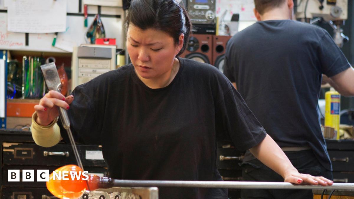 A woman creates a jar at a glass-blowing factory. The jar is glowing red at the end of the pole used to create it.