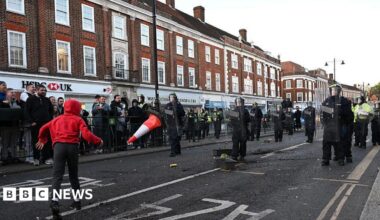 Police are seen on Epsom high street as people come out to protest after a woman was raped last Saturday on April 15, 2026 in Epsom. Someone in a red hoodie throws a traffic cone at police.