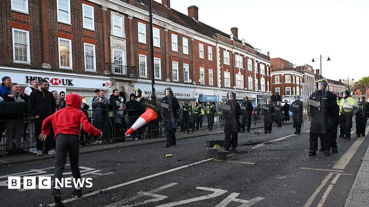 Police are seen on Epsom high street as people come out to protest after a woman was raped last Saturday on April 15, 2026 in Epsom. Someone in a red hoodie throws a traffic cone at police.