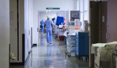 A stock image of a hospital corridor, with two health workers in sanitary clothing walking away from the camera