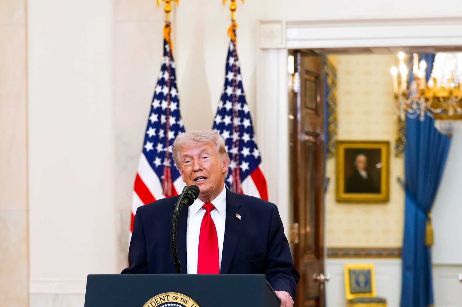 Donald Trump speaking at a podium bearing the presidential seal. Behind him, somewhat blurred, are a red carpet, two furled American flags and what looks like a room in the White House.