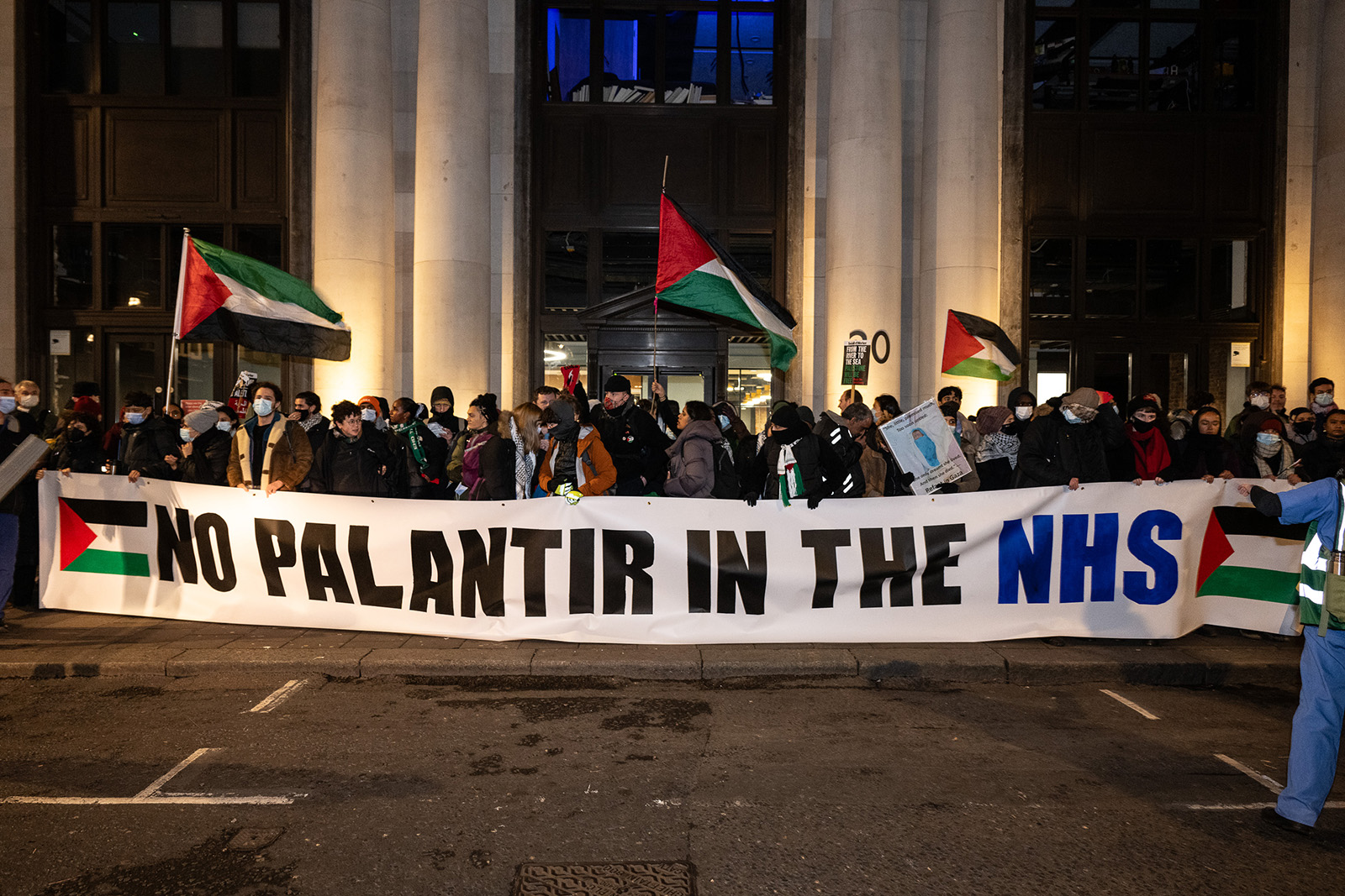 A photograph of a Health Workers for a Free Palestine protest outside Palantir's London office in December 2023. The demonstrators are holding a 'No Palantir in the NHS' banner. 