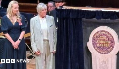 Lissie Harper and Geraldine Winner, who has short, white hair and wears a loose, grey suit, stand on a wooden stage next to a stone memorial in Andrew Harper's name. Curtains have been opened on either side to reveal the memorial.