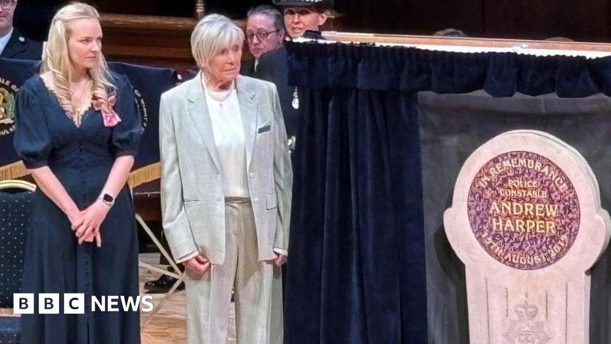Lissie Harper and Geraldine Winner, who has short, white hair and wears a loose, grey suit, stand on a wooden stage next to a stone memorial in Andrew Harper's name. Curtains have been opened on either side to reveal the memorial.
