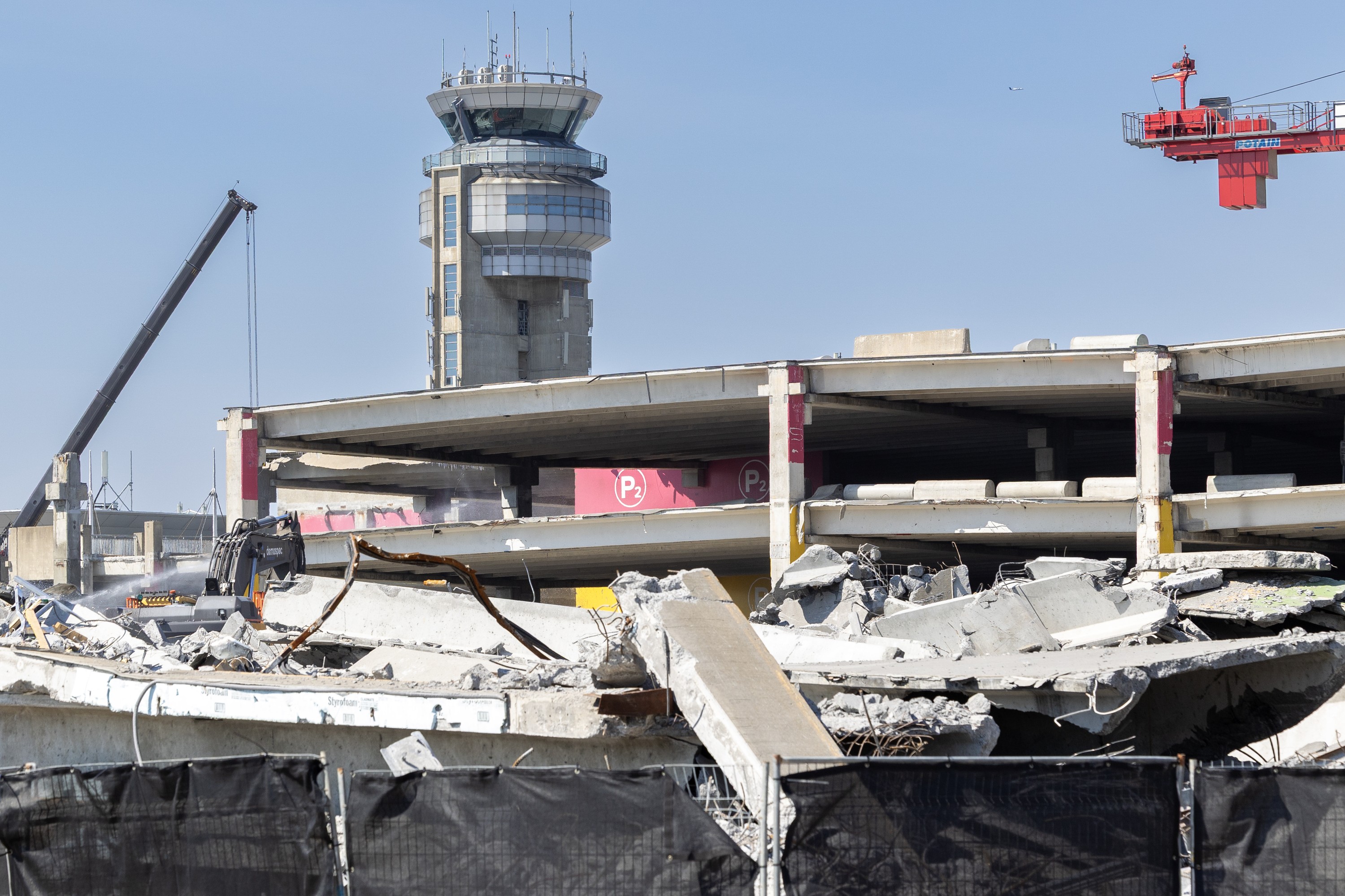 An airport control tower is seen behind a parking garage in the process of being demolished.