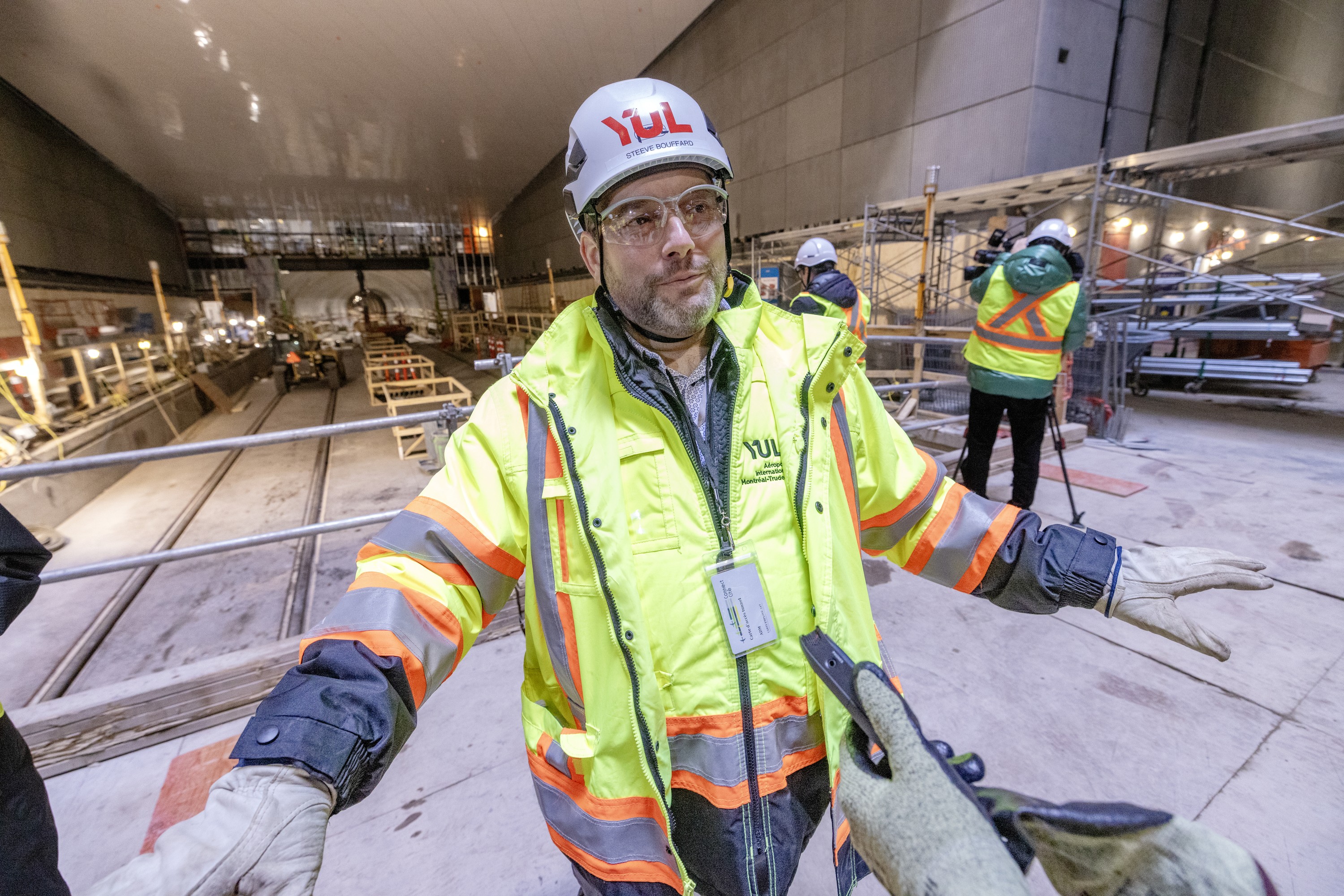 A man in construction gear speaks on a platform in a large tunnel.