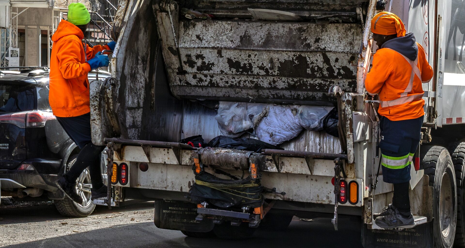 Two people in orange jackets on either side of the back of a garbage truck.