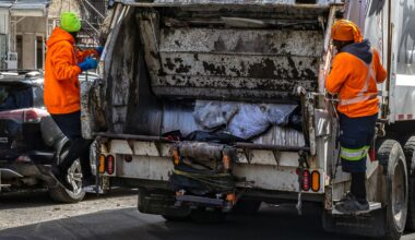 Two people in orange jackets on either side of the back of a garbage truck.