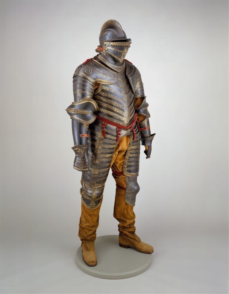 A full suit of medieval armor displayed on a stand, featuring ornate detailing and paired with suede-like boots, in a museum setting