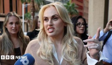 A blonde haired woman speaks into a microphone on a street with people standing behind her