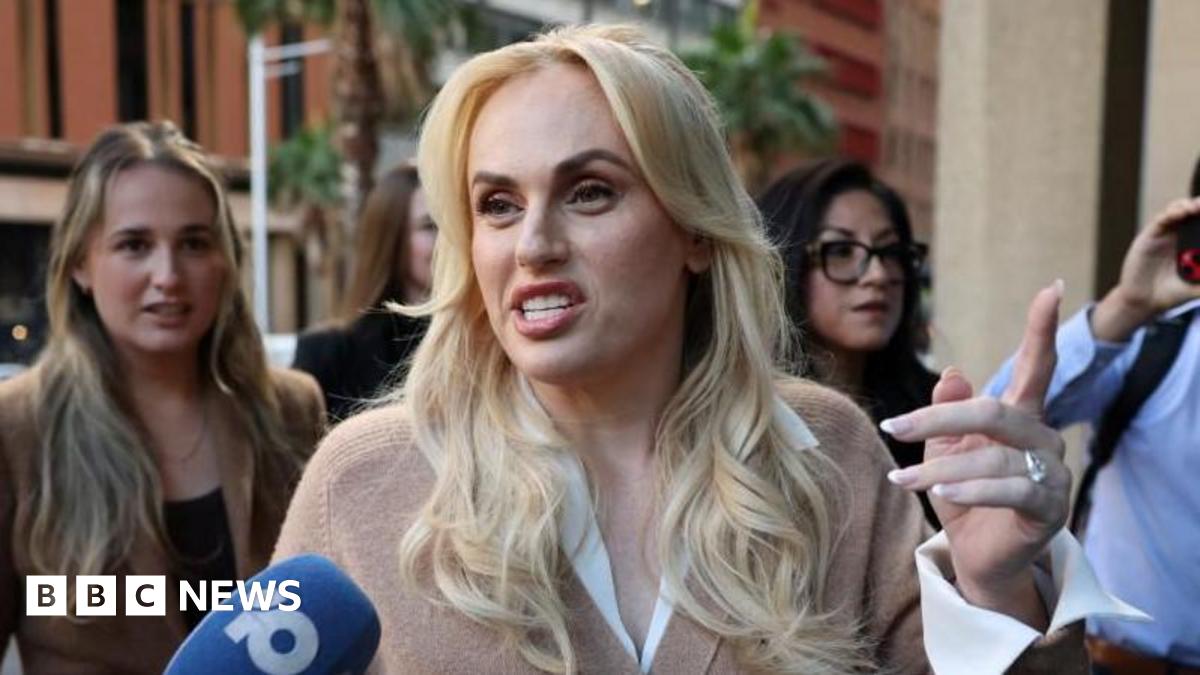 A blonde haired woman speaks into a microphone on a street with people standing behind her
