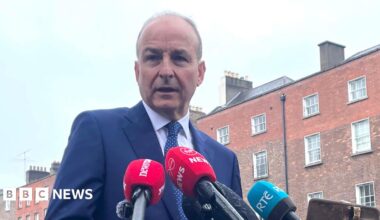 Micheál Martin, a man with short, receding grey hair, speaks into microphones while standing on a street in central Dublin.  He is wearing a navy blue suit and a navy blue polka dot tie. A terrace of tall, red brick Georgian town houses line the road behind him.