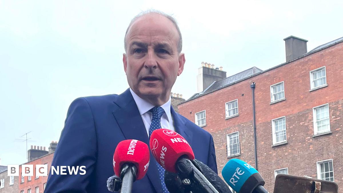 Micheál Martin, a man with short, receding grey hair, speaks into microphones while standing on a street in central Dublin.  He is wearing a navy blue suit and a navy blue polka dot tie. A terrace of tall, red brick Georgian town houses line the road behind him.