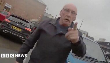 A man standing in a car park. He has his finger pointed at the person wearing the body-cam.