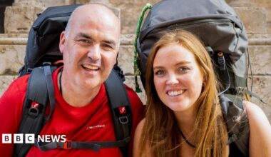 two people looking at the camera - they are a bald man and a red-headed woman. Both are wearing big rucksacks