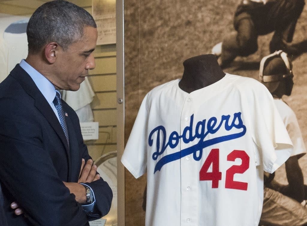 A man in a suit observes a Dodgers jersey with the number 42, displayed in a museum setting, alongside a historical baseball photo