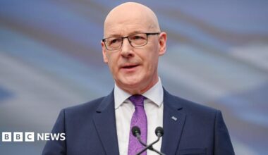 John Swinney, who is bald with glasses, speaks into a microphone. He is wearing a business suit and purple shirt
