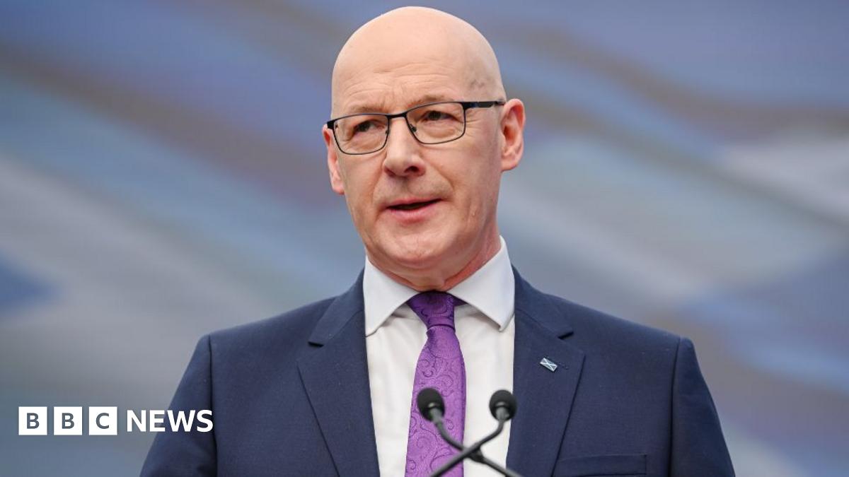 John Swinney, who is bald with glasses, speaks into a microphone. He is wearing a business suit and purple shirt