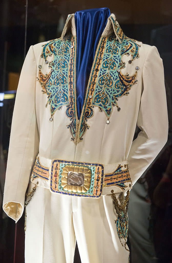 Ornate white jumpsuit with intricate patterns and embellishments, featuring a large decorative belt buckle, displayed on a mannequin