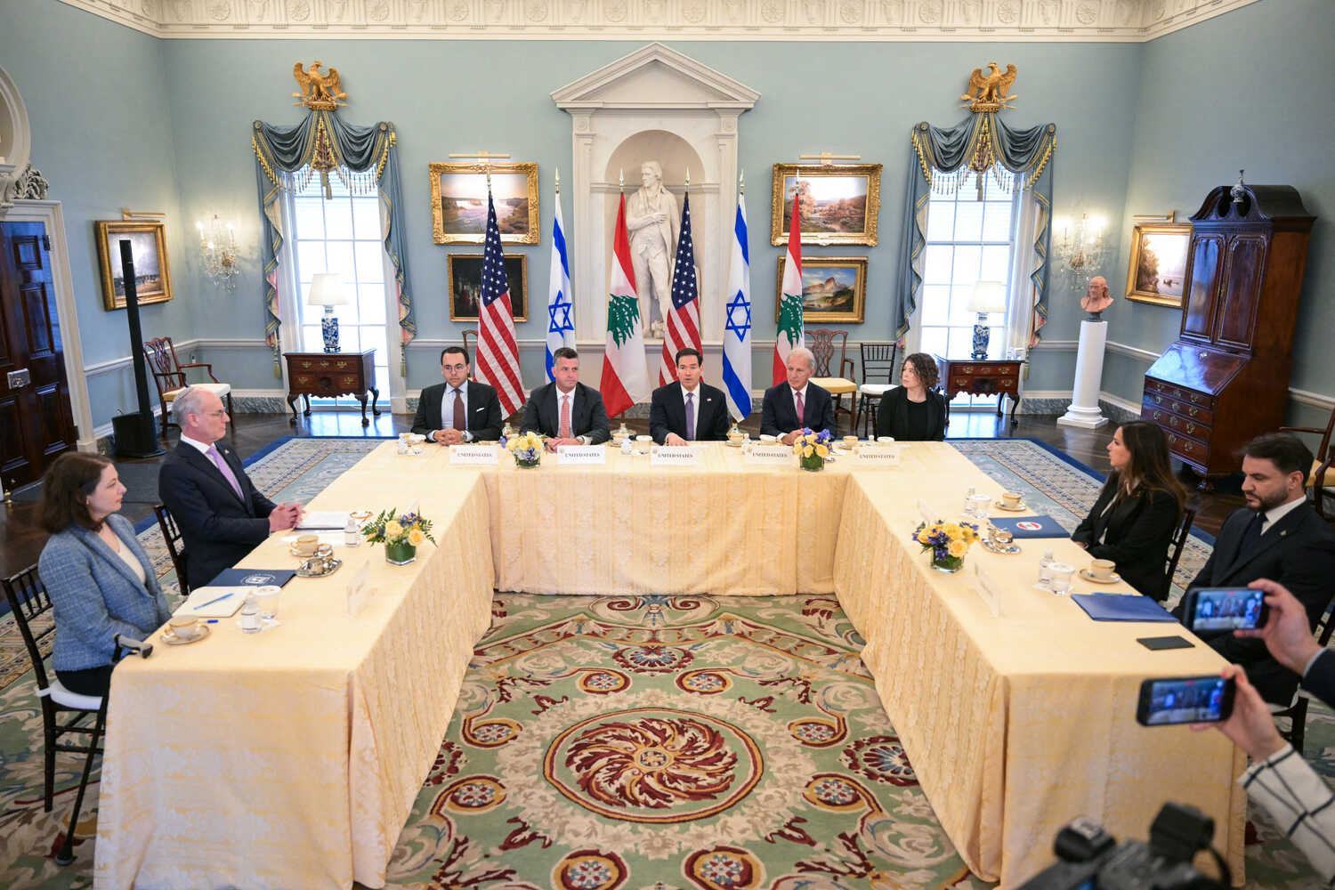 People are seated around a large U-shaped table in front of American, Israeli, and Lebanese flags.