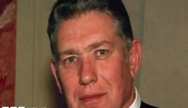 A man with short dark hair looking ahead. He is wearing a white shirt and black jacket.