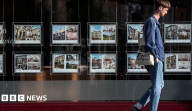 A man wearing blue jeans and a blue shirt walks past an estate agent window displaying home listing photos.