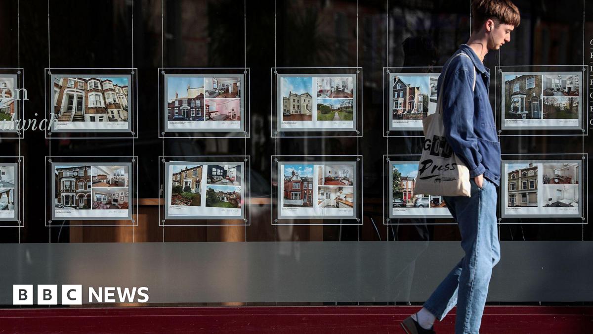 A man wearing blue jeans and a blue shirt walks past an estate agent window displaying home listing photos.