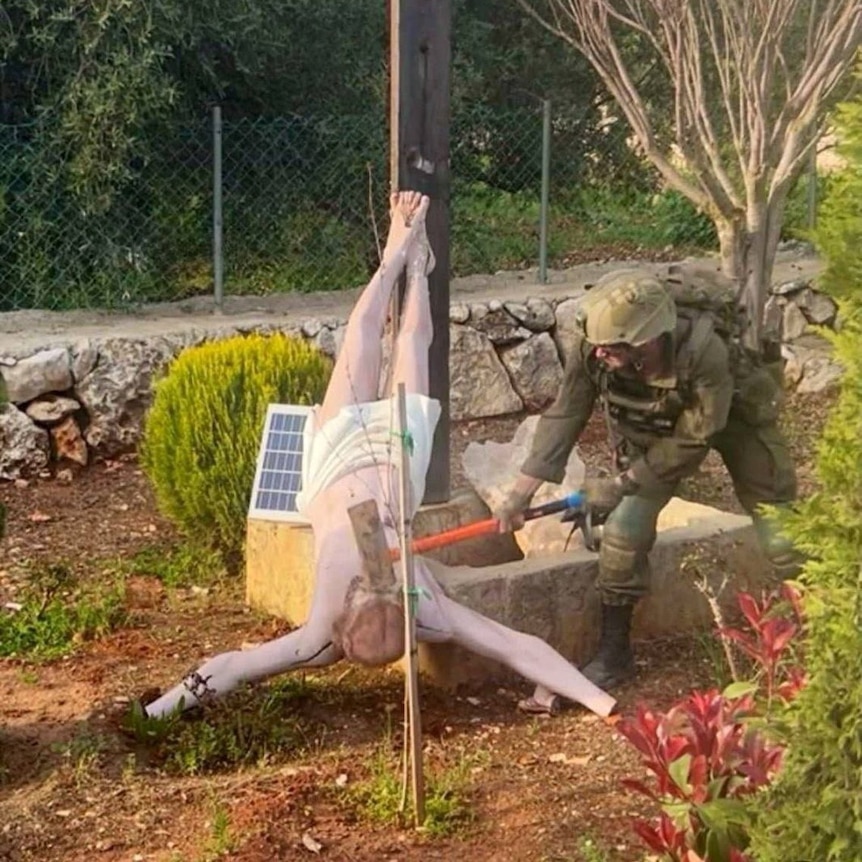 A soldier in uniform swings the blunt side of an axe at a fallen sculpture of Jesus on a corss