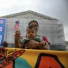 Olga Urbina holds her 9-month-old son, Ares Webster, in a front-pack baby carrier at a protest in front of the Supreme Court building. Ares holds a small American flag. Another protester holds a sign that says, "American Born Children Are American Children."