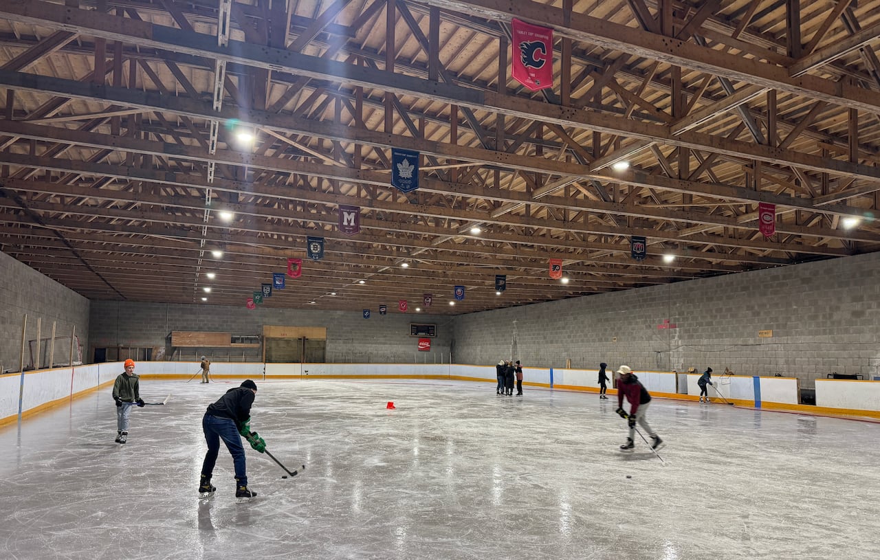 People skate and play hockey on an ice rink inside a grey cinderblock building with wooden rafters.