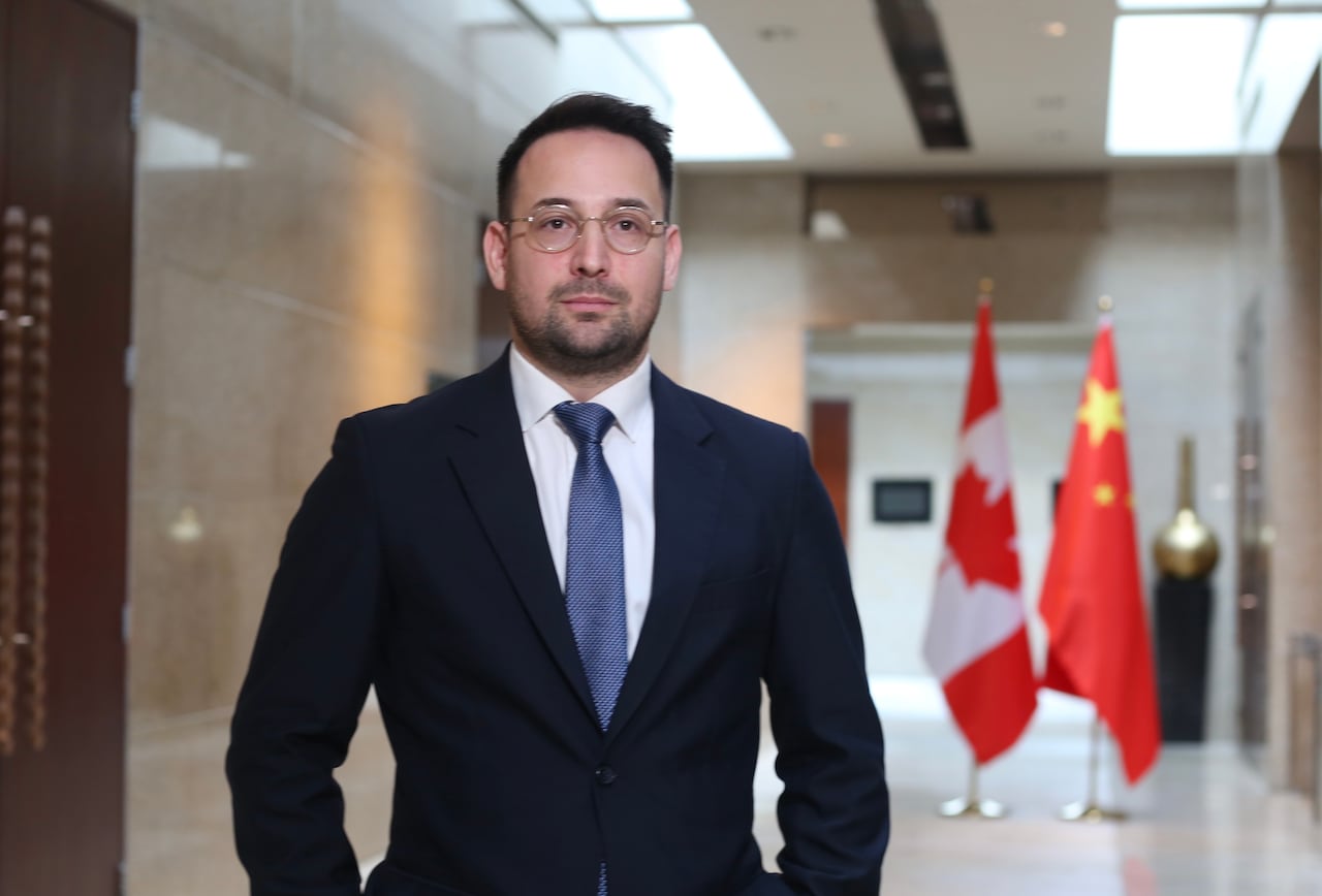 A man in a dark suit and glasses stands in a hallway.