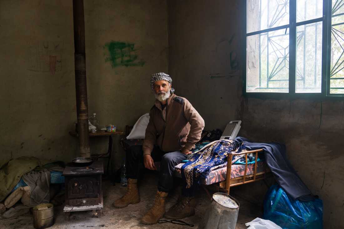 Joseph Elias Issa, 56, sits on a day bed next to a wood burning stove in a small cement building. He has a white beard and a scarf wrapped on his head.