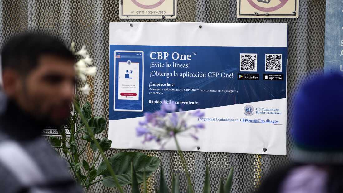 People waiting to apply for asylum stand in front of a sign for the CBP One app as they camp near the pedestrian entrance to the San Isidro Port of Entry, linking Tijuana, Mexico with San Diego.