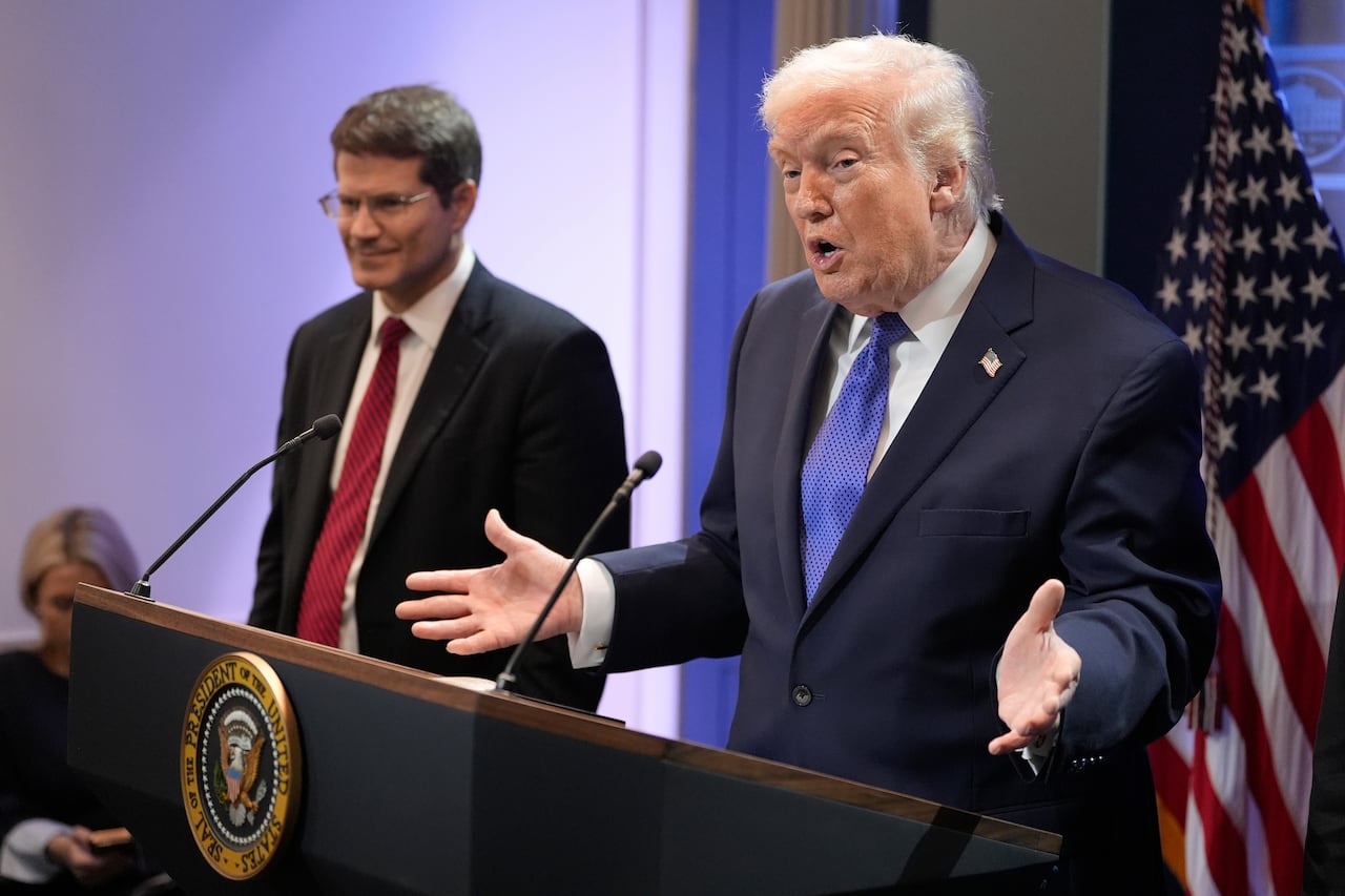 A cleanshaven man speaks at a podium as another man, bespectacled, looks on. An American flag is also shown. 