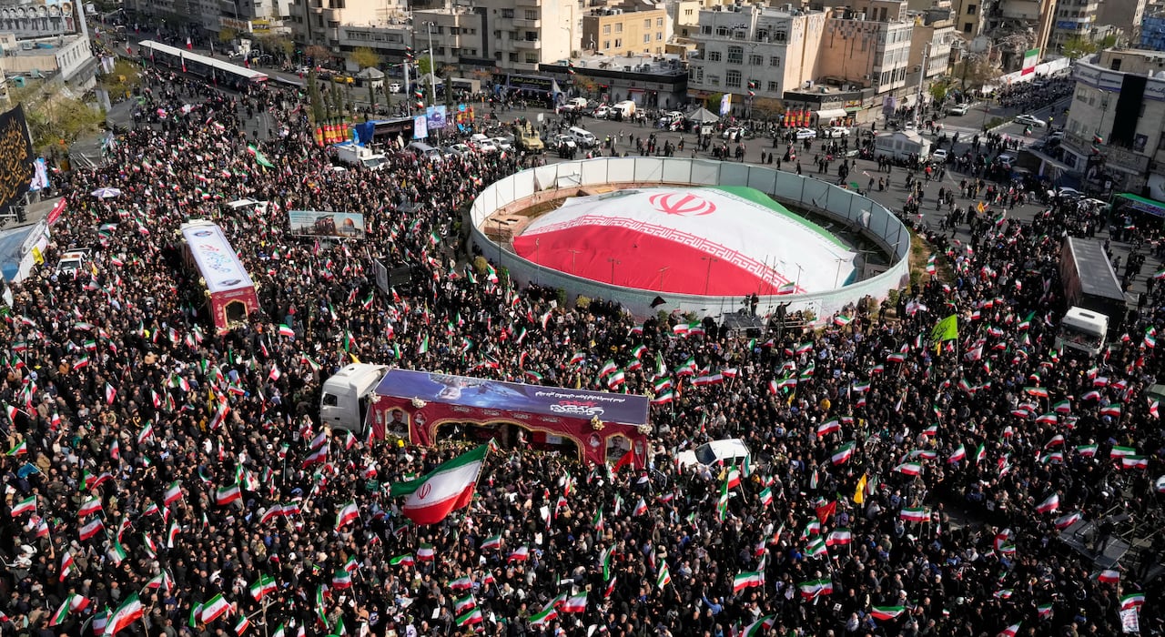 A large crowd in the streets encircling a giant Iranian flag, viewed from above.  