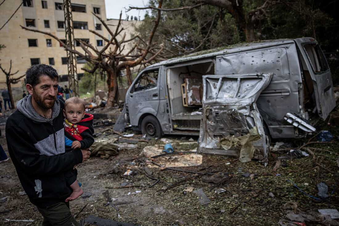 A man carries his child as he walks past destroyed vehicles and debris at the site of an overnight Israeli airstrike that killed seven people on April 01, 2026 in Beirut, Lebanon.
