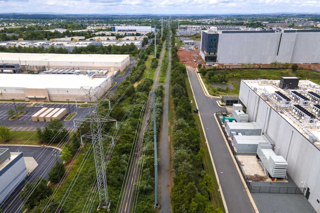 High-voltage transmission lines extend along a grassy green strip of land, providing electricity to large, box-shaped data centers that sit on both sides of the strip in Ashburn, Virginia.