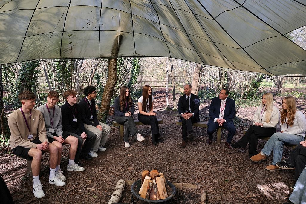 People sit on benches in a circle under a covering with a firepit in the middle.